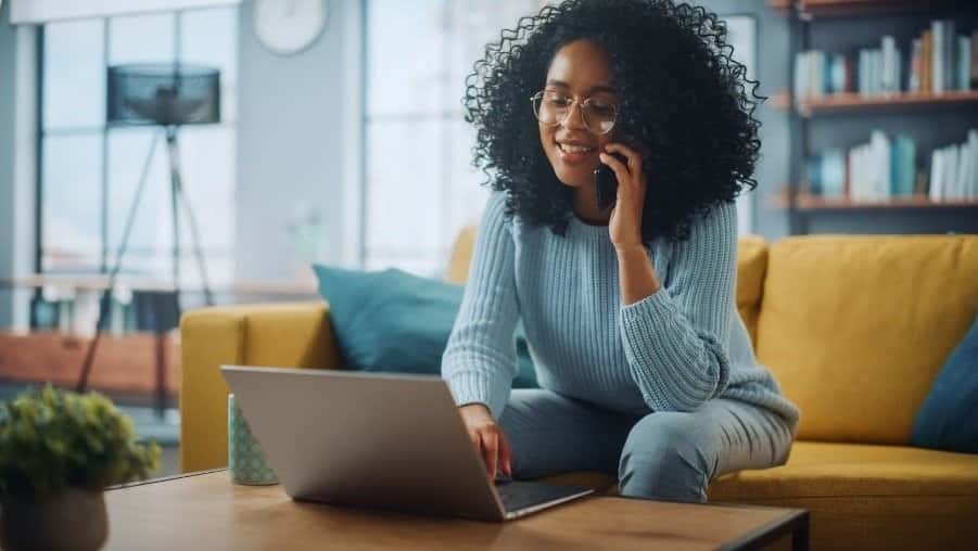 A woman sitting down on her sofa. She is on the smiling whilst on a call and using her laptop